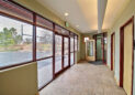 A building entrance features glass doors and large windows overlooking a parking lot and trees. The tiled floor leads to several wooden doors, one with a restroom sign. Natural light floods the space, highlighting the neutral walls.