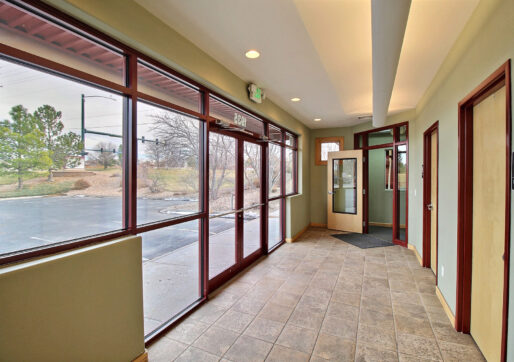 A building entrance features glass doors and large windows overlooking a parking lot and trees. The tiled floor leads to several wooden doors, one with a restroom sign. Natural light floods the space, highlighting the neutral walls.