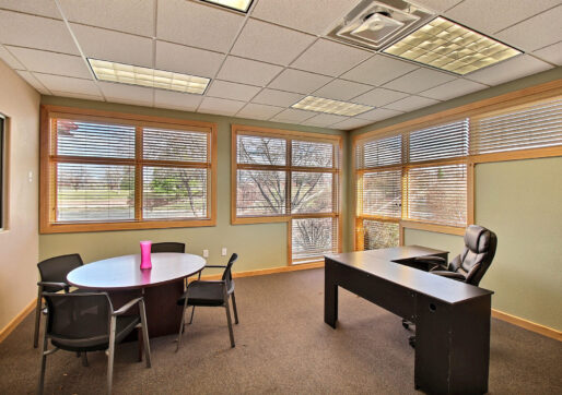 A bright office with large windows, featuring a wooden desk with a black leather chair and a round table with three chairs. A pink water bottle is on the table. The room has beige walls and carpeted flooring.