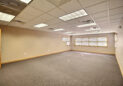 An empty office room with beige walls, a grey carpet, and a suspended ceiling with fluorescent lights. Large windows on one wall allow natural light, and a wooden door is slightly open.