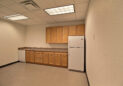 A small, empty kitchenette with light wood cabinets, a countertop, and a white fridge. There is also a dishwasher and a sink. The flooring is linoleum, and the room has a drop ceiling with fluorescent lights.