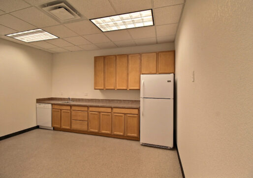 A small, empty kitchenette with light wood cabinets, a countertop, and a white fridge. There is also a dishwasher and a sink. The flooring is linoleum, and the room has a drop ceiling with fluorescent lights.