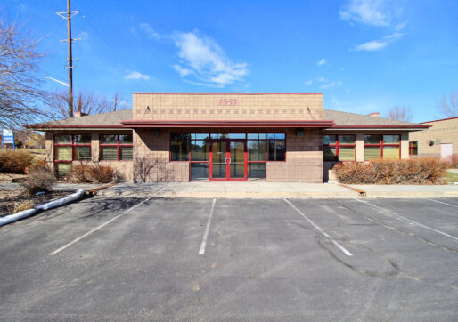 A single-story commercial building with large windows and a brick facade marked "1935" above the entrance. The building is surrounded by a paved parking area with some empty parking spaces. The sky is clear and blue.