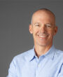 A smiling man with a shaved head is wearing a light blue button-up shirt. He stands against a plain gray background with his arms casually crossed.