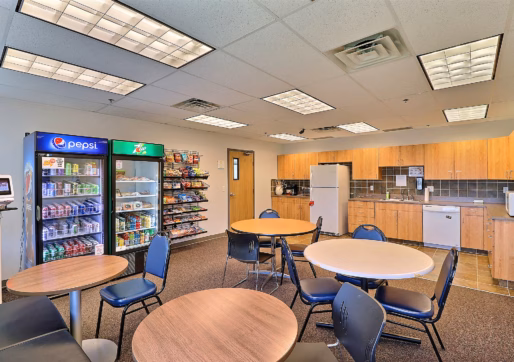 A break room with round tables and chairs, vending machines, snack racks, refrigerator, microwave, sink, coffee maker, and wooden cabinets under bright fluorescent lighting—ideal for tenants of the Office/Medical Suite For Lease.