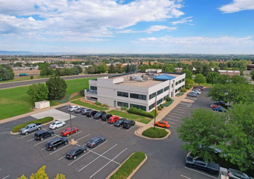 Aerial view of a modern, two-story office building with an Office/Medical Suite For Lease, a parking lot surrounded by trees, several parked cars, and a grassy field in the background under a partly cloudy sky.