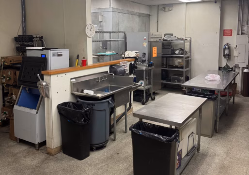 A commercial kitchen with stainless steel counters, trash bins, shelves with equipment, a clock on the wall, and overhead fluorescent lights. The space appears clean and organized, with various appliances and utensils visible.