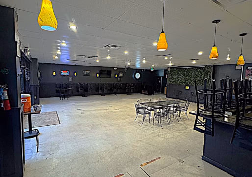 Spacious, empty dining area with hanging yellow lights, stacked chairs on the right, and a few tables in the center. The walls are dark, and the room appears to be closed or preparing to open.