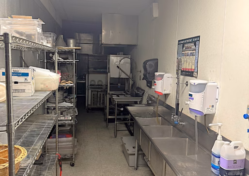 A commercial kitchen with metal shelves holding food containers on the left and stainless steel sinks on the right. Soap dispensers and cleaning supplies are above the sinks; a dishwasher and kitchen equipment are visible at the back.