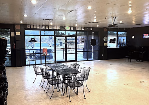 A spacious, mostly empty room with a set of black wire chairs and tables in the center, large glass windows and doors at the far end, and a TV mounted on the wall to the right. Light shines in from outside.