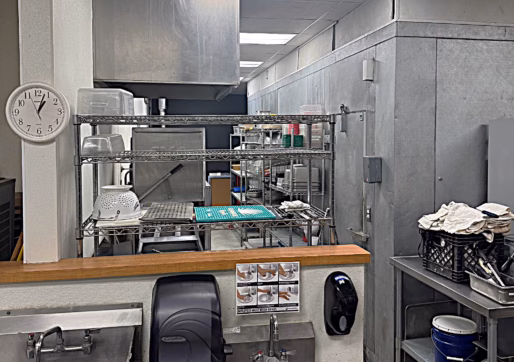 A commercial kitchen dishwashing area with a metal sink, soap and paper towel dispensers, a wall clock, shelves with dishes and plastic bins, and stainless steel refrigerators under fluorescent lighting.