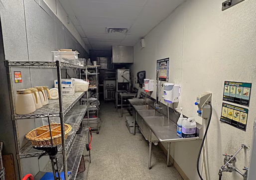A commercial kitchen dishwashing area with metal shelves holding dishes and bowls on the left, and stainless steel sinks, soap dispensers, and cleaning supplies on the right. The room is brightly lit with fluorescent lights.