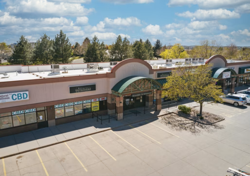 Space For Lease in a neighborhood shopping mall with several storefronts, under a blue sky with scattered clouds. There are a few parked cars and trees with green leaves in the parking lot.