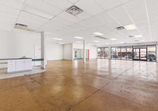 Retail For Lease-4835 W 10th St, Unit A, Greeley, CO 80634 - A bright, empty commercial space with large front windows, white walls, a brown polished floor, ceiling tiles with fluorescent lighting, and a small counter area on the left side near a door.