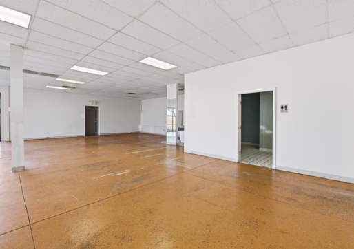 Retail For Lease-4835 W 10th St, Unit A, Greeley, CO 80634 -A spacious commercial room with brown concrete floors, white walls, and a drop ceiling with fluorescent lights. Two doors and windows are visible, and one room has a tiled floor.