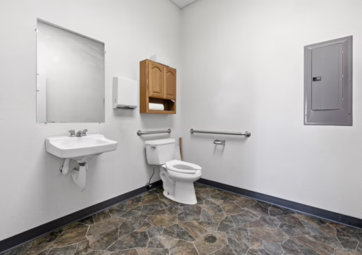 Retail For Lease-4835 W 10th St, Unit A, Greeley, CO - A clean, accessible bathroom with a toilet, grab bars, sink, mirror, paper towel dispenser, wooden cabinet, and a gray wall-mounted electrical panel. The floor has a patterned tile design.