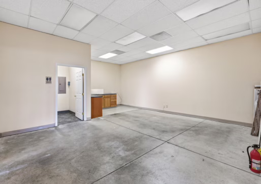 Retail For Lease-4835 W 10th St, Unit A, Greeley, CO 80634 - An empty room with beige walls, exposed concrete floor, drop ceiling with fluorescent lights, a doorway leading to another area, a small counter with a sink, and a red fire extinguisher in the corner.
