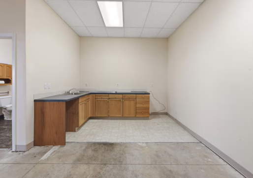 Retail For Lease-4835 W 10th St, Unit A, Greeley, CO 80634 - A kitchenette with wood cabinets, a countertop, and a sink. The floor is partly unfinished, and a bathroom with visible toilet and vanity is seen through an open door on the left.