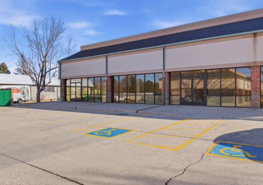 Retail For Lease-4835 W 10th St, Unit A, Greeley, CO 80634 - A strip mall with large glass windows and doors, an empty parking lot, and two clearly marked handicap parking spaces in front. A tree is visible to the left.