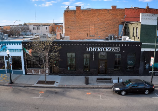 For Sale-909 8th Ave, Greeley, CO 80631-A black-painted building with a sign reading “PATRICK’S IRISH PUB” sits on a city street, with a parked car and a tree in front. This retail bar for sale is surrounded by adjacent buildings of various colors and styles.
