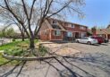 For Lease-919 44th Ave Ct, Greeley, CO. A two-story brown townhouse with white trim sits on a corner lot. Leafless trees and landscaped garden beds are in front. Several cars are parked in driveways on a sunny day with a clear blue sky.