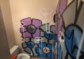 Office For Lease-809 9th St, Unit 2A, Greeley, CO 80631 - A small bathroom with tan walls covered in colorful graffiti-style flower murals in shades of blue, purple, and white. There is a toilet, a vent on the floor, and a vent on the ceiling.