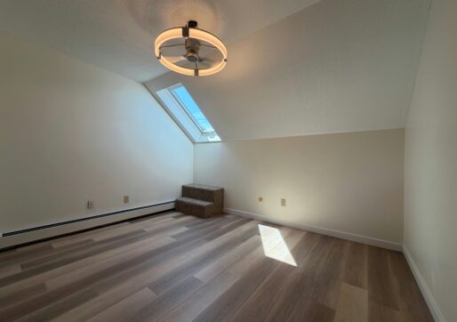 For Lease-919 44th Ave Ct, Unit D, Greeley, CO 80634 - Empty attic room with sloped ceiling, large skylight, modern ceiling light, and small carpeted steps by the window. Sunlight streams onto the wooden floor, creating a bright and airy atmosphere in this stylish townhome for rent.