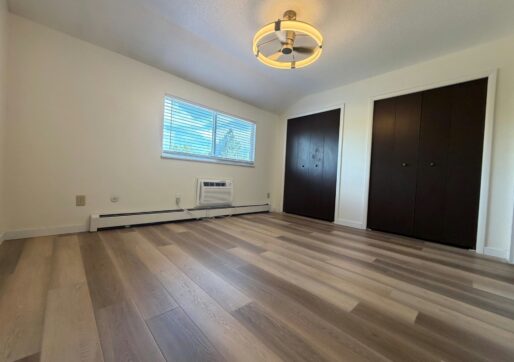 For Lease-919 44th Ave Ct, Unit D, Greeley, CO 80634 - A bright, empty room in a townhome for rent features light wood flooring, a modern ceiling light, a window with blinds, an air conditioning unit, and two dark-colored closet doors.