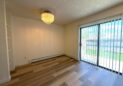 For Lease-919 44th Ave Ct, Unit D, Greeley, CO. A bright, empty room with light wood flooring, a modern chandelier, white walls, built-in shelves, and large sliding glass doors with vertical blinds leading to a grassy fenced backyard.