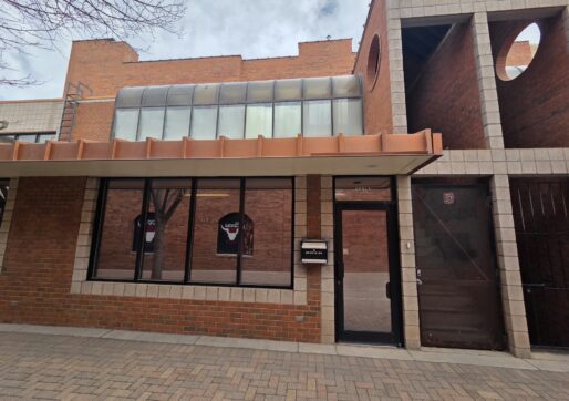 Office For Lease-809 9th St, Unit 2A, Greeley, CO 80631 - A brick building with large windows, a mailbox labeled "mail drop," and a glass door. The building features tan trim and a covered walkway with a brick-paved sidewalk in front.