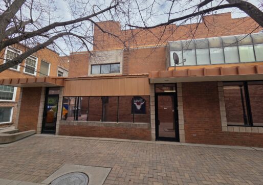 Office For Lease-809 9th St, Unit 2A, Greeley, CO 80631 - A brick building with large windows and a brown awning, bordered by leafless tree branches. The sidewalk is paved with bricks, and there is a glass door and satellite dish on the right side of the building.