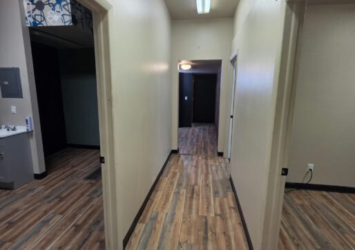 Office For Lease-809 9th St, Unit 2A, Greeley, CO 80631 - A hallway with light-colored walls and wood-patterned floors, featuring several open doorways on both sides. Fluorescent lights are on the ceiling, and rooms are visible through the doorways.