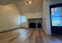 For Lease-919 44th Ave Ct, Unit D, Greeley, CO. A bright, empty living room with light wood flooring, high vaulted ceiling, white walls, a brick fireplace, a window with blinds, and a glass door leading outside.