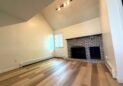 For Lease-919 44th Ave Ct, Unit D, Greeley, CO. A bright, empty living room with light wood floors, white walls, a high vaulted ceiling, a window with blinds, and a brick fireplace with two black metal covers. Ceiling track lighting is turned on.