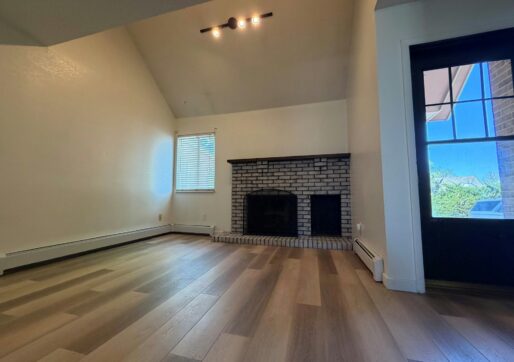 For Lease-919 44th Ave Ct, Unit D, Greeley, CO. A bright, empty living room with light wood flooring, high vaulted ceiling, white walls, a brick fireplace, a window with blinds, and a glass door leading outside.