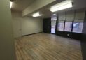 Office For Lease-809 9th St, Unit 2A, Greeley, CO 80631 - An empty room with wood-style flooring, white and dark walls, a closed door, fluorescent ceiling lights, and large windows facing a brick building outside.