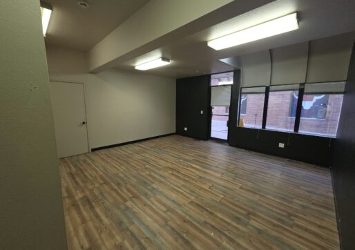 Office For Lease-809 9th St, Unit 2A, Greeley, CO 80631 - An empty room with wood-style flooring, white and dark walls, a closed door, fluorescent ceiling lights, and large windows facing a brick building outside.
