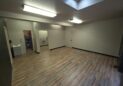 Office For Lease-809 9th St, Unit 2A, Greeley, CO 80631 - A spacious, empty room with wood-style flooring, beige walls, fluorescent ceiling lights, a skylight, open doorways leading to other rooms, and a small sink in one corner.