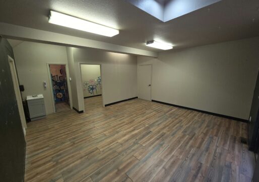 Office For Lease-809 9th St, Unit 2A, Greeley, CO 80631 - A spacious, empty room with wood-style flooring, beige walls, fluorescent ceiling lights, a skylight, open doorways leading to other rooms, and a small sink in one corner.