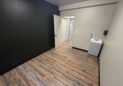 Office For Lease-809 9th St, Unit 2A, Greeley, CO 80631 - Empty room with wood-patterned flooring, one black wall, beige walls, an open door leading to a hallway, and a small gray sink with a soap dispenser on the right side.
