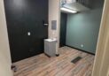 Office For Lease-809 9th St, Unit 2A, Greeley, CO 80631 - A small, empty room with dark green walls and wood-patterned flooring. A utility sink and paper towel dispenser are mounted against one wall. Overhead fluorescent lighting brightens the space.