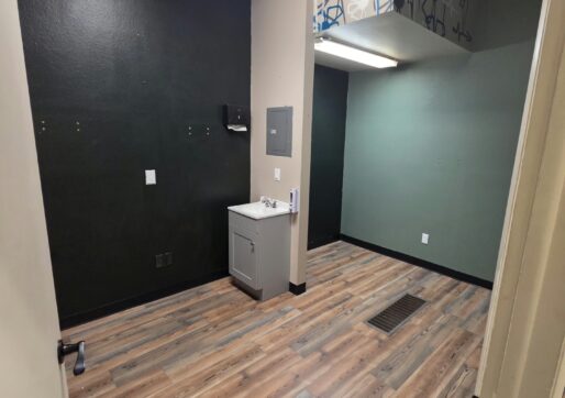 Office For Lease-809 9th St, Unit 2A, Greeley, CO 80631 - A small, empty room with dark green walls and wood-patterned flooring. A utility sink and paper towel dispenser are mounted against one wall. Overhead fluorescent lighting brightens the space.