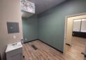 Office For Lease-809 9th St, Unit 2A, Greeley, CO 80631 - A small room with green walls, wood-patterned flooring, a gray cabinet with a white sink, a wall vent, and a ceiling with a decorative floral panel. An open doorway leads to another room with similar flooring.