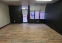 Office For Lease-809 9th St, Unit 2A, Greeley, CO 80631 - An empty room with dark walls, large windows, wooden flooring, and fluorescent lights on the ceiling. A door and a Texas flag can be seen through the windows.