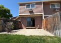 For Lease-919 44th Ave Ct, Unit D, Greeley, CO 80634 - A fenced backyard with a small concrete patio, patch of grass, and a two-story brown townhome for rent. The sliding glass door and windows face the yard; a bush and gravel line the fence on the left.