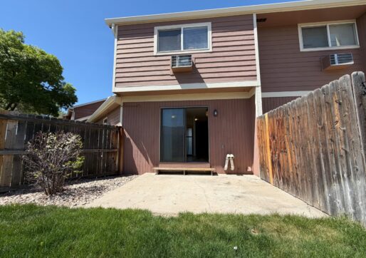 For Lease-919 44th Ave Ct, Unit D, Greeley, CO 80634 - A fenced backyard with a small concrete patio, patch of grass, and a two-story brown townhome for rent. The sliding glass door and windows face the yard; a bush and gravel line the fence on the left.