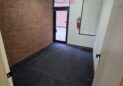 Office For Lease-809 9th St, Unit 2A, Greeley, CO 80631 - A small, empty office room with dark carpet, a brick wall on one side, and a glass door leading outside. There is a bright ceiling light, an emergency exit sign, and a window covered by a white blind.