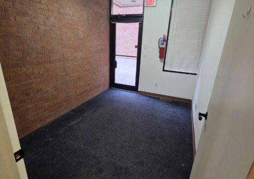 Office For Lease-809 9th St, Unit 2A, Greeley, CO 80631 - A small, empty office room with dark carpet, a brick wall on one side, and a glass door leading outside. There is a bright ceiling light, an emergency exit sign, and a window covered by a white blind.