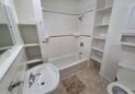 For Sale or Lease-222 Oak Ave, Eaton, CO 80615 - A small bathroom with a white bathtub and shower, white sink, open shelves, a toilet, and tiled floor. The walls are white with some decorative tile accents above the tub.