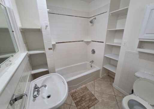 For Sale or Lease-222 Oak Ave, Eaton, CO 80615 - A small bathroom with a white bathtub and shower, white sink, open shelves, a toilet, and tiled floor. The walls are white with some decorative tile accents above the tub.